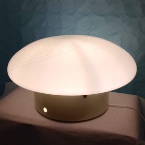 May include: A white mushroom-shaped table lamp with a rounded top and a cylindrical base. The lamp emits a soft glow, and the top features subtle, curved lines. The lamp is on a white surface against a blue background.