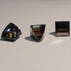 May include: Three faceted gemstones in shades of grey, green, and gold. The gemstones are cut into a triangle, a square, and a modified square shape. The gems are displayed on a white surface.