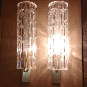 Op de afbeelding: Een paar cilindrische wandlampen met heldere, getextureerde glazen kappen met een verticaal bamboe-achtig patroon. Elk armatuur is gemonteerd op een rechthoekige messingkleurige metalen voet, waarbij één lamp brandt voor licht.