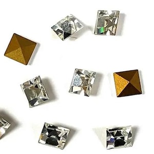 May include: Close-up of several square-shaped rhinestones. Some are clear and faceted, reflecting light, while others are a solid gold color. The rhinestones are scattered on a white background.