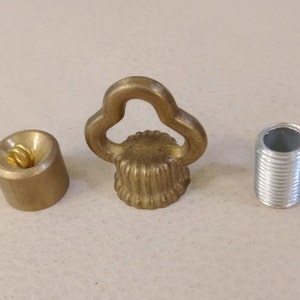 Peut inclure: Trois pièces de quincaillerie en métal : un capuchon de couleur laiton avec une boucle, une pièce cylindrique de couleur laiton avec un ressort à l'intérieur et un cylindre fileté de couleur argent. Les pièces sont sur une surface claire.