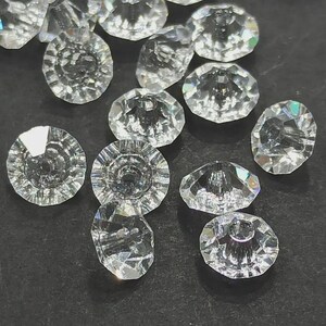 Puede incluir: Un conjunto de cuentas de cristal transparentes y facetadas. Las cuentas tienen forma redonda, similar a un botón, con un agujero central. Las facetas capturan la luz, creando un efecto brillante. Ideal para la creación de joyas o proyectos de artesanía.