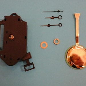 May include: Clock repair parts including a dark brown clock movement, three black clock hands, a gold-colored pendulum, and two small metal pieces. The parts are arranged on a light blue surface.