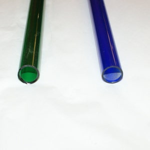 Puede incluir: Dos tubos de vidrio, uno verde y otro azul, se muestran sobre una superficie blanca. Los tubos son transparentes y tienen un acabado liso y pulido.