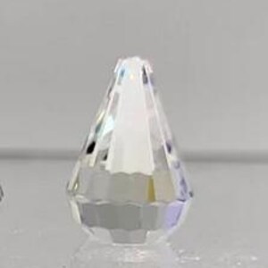 Könnte beinhalten: Eine klare, facettierte Kristallperle in Tropfenform. Die Perle hat eine spitze Spitze und eine flache Basis, die das Licht reflektiert und einen funkelnden Effekt erzeugt. Der Kristall ist vor einem neutralen Hintergrund platziert.