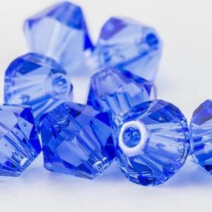 Könnte beinhalten: Nahaufnahme von mehreren facettierten, transparenten blauen Perlen. Die Perlen sind tiefblau und haben ein geometrisches Design mit mehreren Facetten. Diese Perlen werden wahrscheinlich für die Schmuckherstellung oder andere Bastelprojekte verwendet.