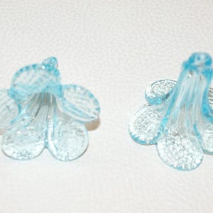 May include: Two light blue, translucent flower-shaped glass beads. Each bead has five petals and a long, slender stem. The beads are lying on a white surface.