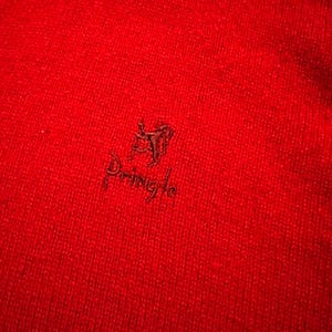 May include: A close-up shot of a bright red knitted textile. The fabric's texture is evident, with a small, dark emblem and the word "Pringle" embroidered on the material. The image suggests a warm, textured item of clothing.