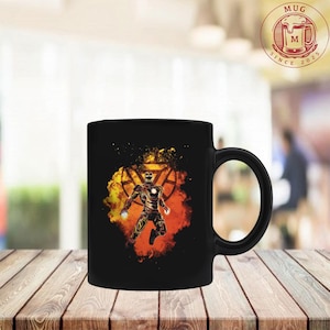 May include: A black ceramic mug featuring a graphic of a robotic figure in a dynamic pose, surrounded by a vibrant orange and yellow explosion effect. The design includes a circular arc reactor symbol behind the figure, set against a dark background.