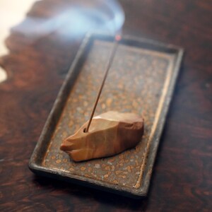 以下が含まれることがあります： 彫刻された石のデザインの茶色とベージュのお香立てで、火のついたお香スティックを持っています。ホルダーは、まだらの模様の長方形のトレイに置かれています。お香から煙が立ち上り、落ち着いた雰囲気を作り出しています。