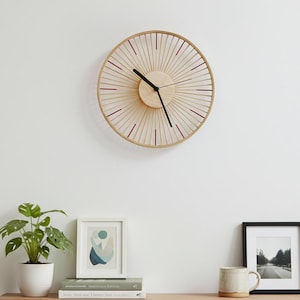 Peut inclure: Une horloge murale ronde en bois clair avec des aiguilles noires. Le cadran présente un motif en forme de soleil avec de fines touches rouges. L'horloge est fixée sur un mur blanc au-dessus d'une étagère en bois.
