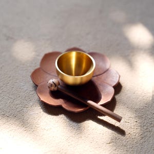 May include: A small, gold singing bowl sits on a brown, flower-shaped base. A wooden mallet with a patterned head lies beside the bowl. The bowl and mallet are designed for sound healing and meditation.