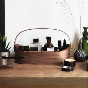 May include: A wooden organizer with a handle, holding various glass bottles and containers. The bottles are filled with liquids and oils. A small black candle and a decorative vase are also present, creating a spa-like atmosphere.