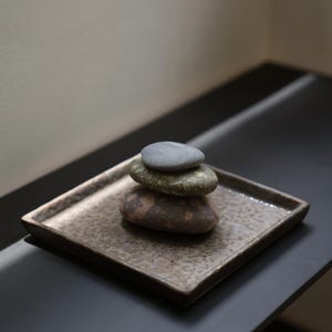 以下が含まれることがあります： 茶色とベージュの斑点模様がある正方形のテクスチャ付きセラミックトレイの上に、滑らかで丸みを帯びた川石が3つ積み重ねられています。石は垂直に配置されており、一番上に明るい灰色の石、真ん中に緑色の石、一番下に茶色の石が置かれています。