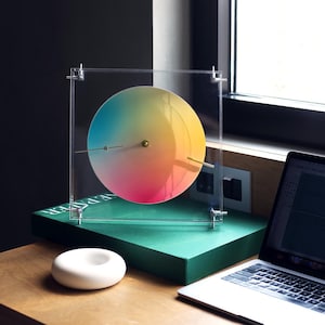 May include: A contemporary clock featuring a circular face with a vibrant rainbow gradient. The clock's hands are gold-toned, and it's housed within a clear acrylic frame. It rests on a green box, with a white ceramic object in the foreground.