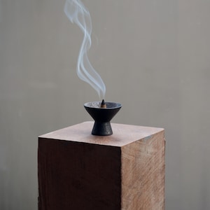 Może przedstawiać: Czarny ceramiczny uchwyt na kadzidło z kadzidłem w kształcie stożka emitującym biały dym. Uchwyt stoi na kwadratowym drewnianym bloku z naturalnym wykończeniem słojów drewna. Tło jest neutralnie szare.