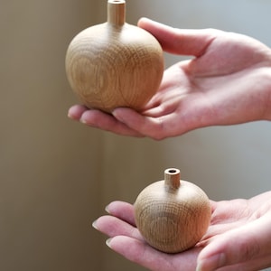 Puede incluir: Dos peque&ntilde;os jarrones redondos de madera con un acabado natural de vetas. Cada jarr&oacute;n tiene un cuello corto y cil&iacute;ndrico. Los jarrones se sostienen en manos abiertas, mostrando su tama&ntilde;o y forma. El fondo es de un tono suave y neutro.