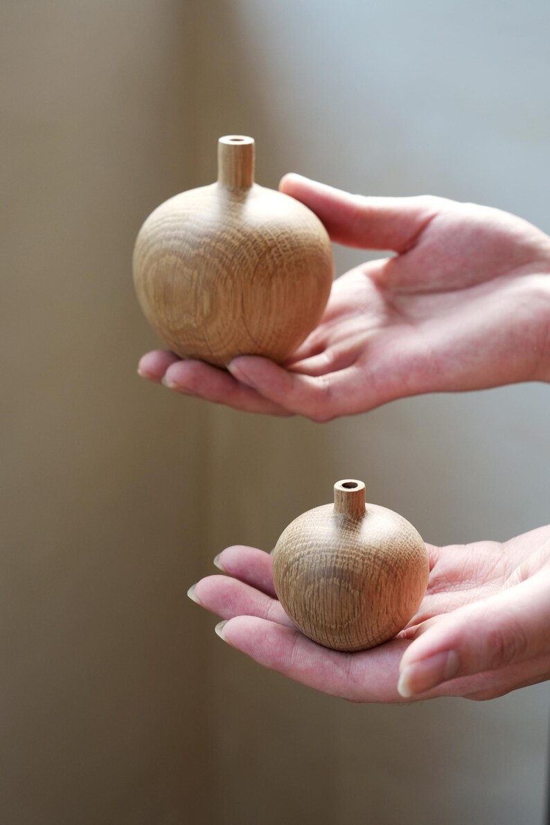 Puede incluir: Dos peque&ntilde;os jarrones redondos de madera con un acabado natural de vetas. Cada jarr&oacute;n tiene un cuello corto y cil&iacute;ndrico. Los jarrones se sostienen en manos abiertas, mostrando su tama&ntilde;o y forma. El fondo es de un tono suave y neutro.
