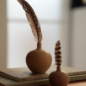 Puede incluir: Dos peque&ntilde;os jarrones de madera con acabado natural, cada uno con una pluma marr&oacute;n y beige. Los jarrones son esf&eacute;ricos con un cuello estrecho. Un jarr&oacute;n est&aacute; enfocado, el otro est&aacute; ligeramente borroso. Los jarrones se asientan sobre una pila de bloques rectangulares de tonos neutros.