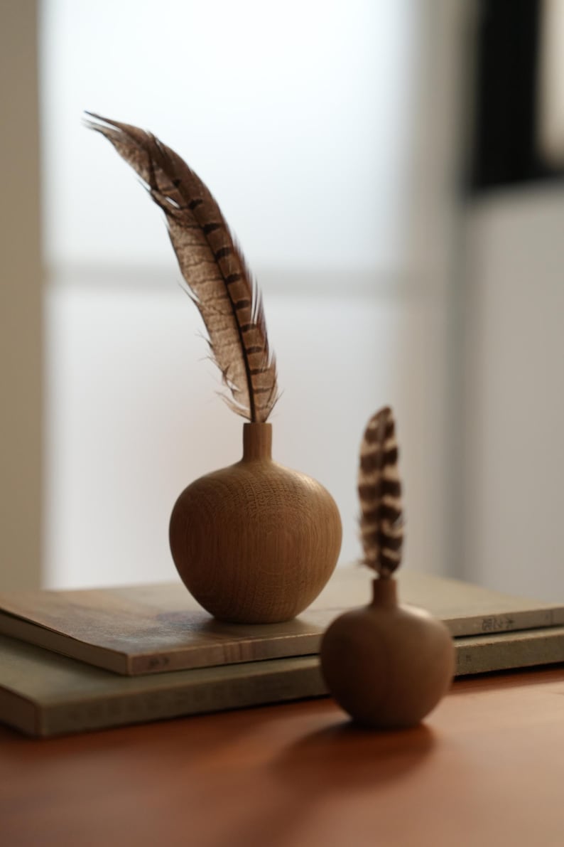 Puede incluir: Dos peque&ntilde;os jarrones de madera con acabado natural, cada uno con una pluma marr&oacute;n y beige. Los jarrones son esf&eacute;ricos con un cuello estrecho. Un jarr&oacute;n est&aacute; enfocado, el otro est&aacute; ligeramente borroso. Los jarrones se asientan sobre una pila de bloques rectangulares de tonos neutros.