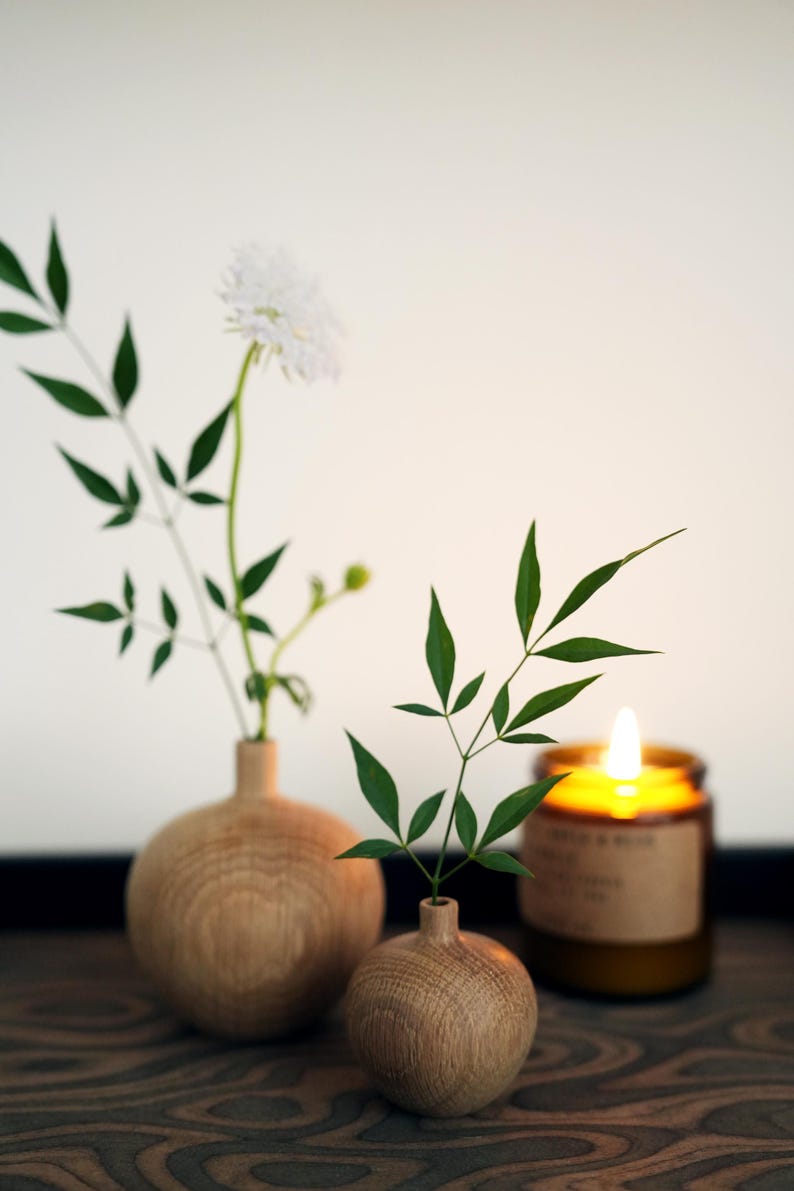 Puede incluir: Dos peque&ntilde;os jarrones redondos de madera sostienen vegetaci&oacute;n y una flor blanca. Una vela encendida en un frasco de vidrio marr&oacute;n est&aacute; en el fondo. Los elementos est&aacute;n dispuestos sobre una superficie de madera oscura, creando una est&eacute;tica natural y minimalista.