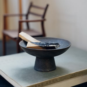 May include: A dark gray ceramic incense bowl with a pedestal base. A piece of wood, partially burned, rests inside the bowl. The bowl sits on a book. A wooden chair is in the background.