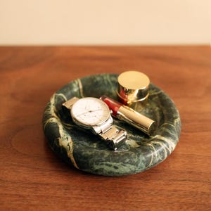 Könnte beinhalten: Ein grünes Marmorschälchen mit einer silbernen Armbanduhr, einem roten Lippenstift und einem kleinen goldfarbenen Behälter. Das runde Schälchen hat ein dunkelgrünes und weißes Adernmuster.