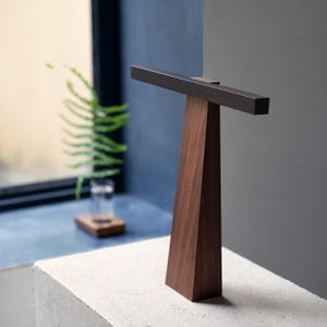 May include: A T-shaped wooden display stand featuring a dark brown horizontal bar resting atop a lighter brown, tapered, triangular vertical base. The structure is crafted from wood with a visible grain, presenting a clean, geometric form.