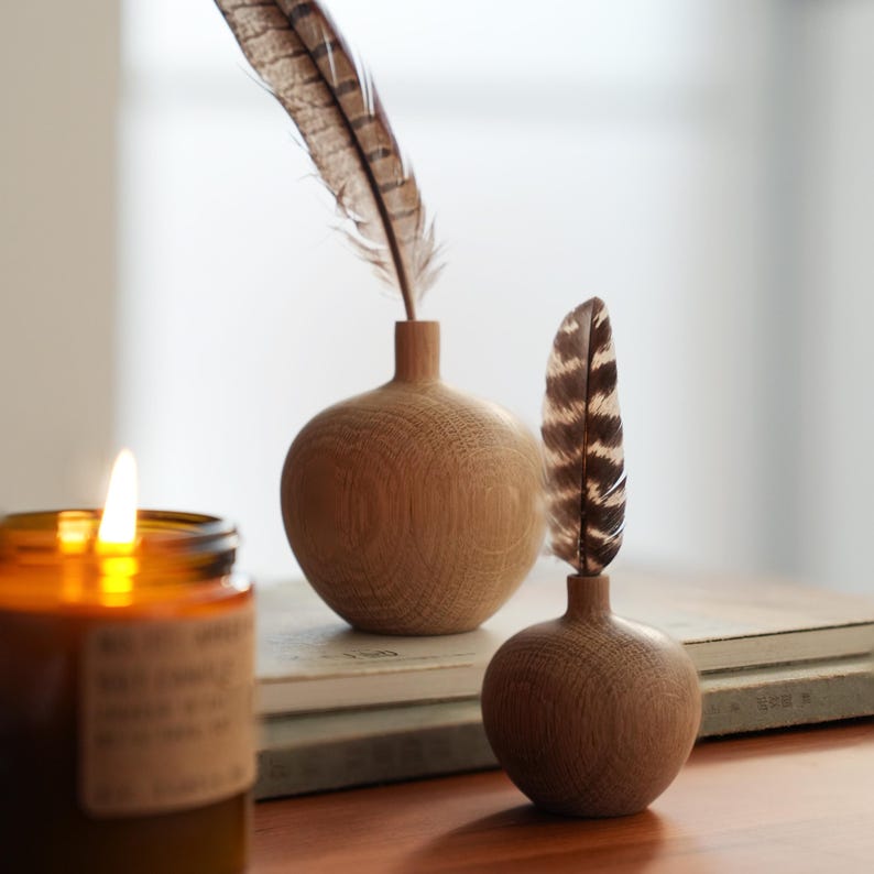 Puede incluir: Dos peque&ntilde;os jarrones redondos de madera con plumas y una vela encendida en un tarro de cristal. Los jarrones son de color marr&oacute;n claro y tienen un dise&ntilde;o sencillo. Las plumas son marrones y blancas. La vela est&aacute; en un tarro de cristal marr&oacute;n.
