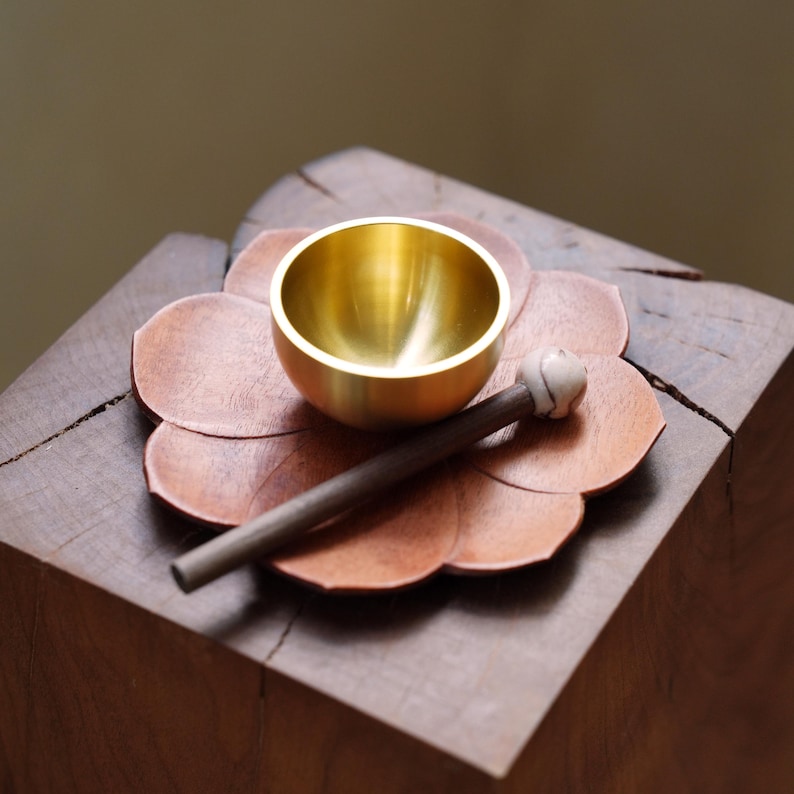 May include: A small, gold-colored singing bowl rests on a copper-colored, flower-shaped base. A wooden mallet with a light-colored, rounded head lies beside the bowl. The objects sit on a dark wooden block, creating a serene, minimalist aesthetic.