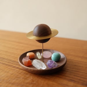 May include: A decorative wooden planet sculpture with a brass ring, mounted on a thin metal rod above a circular wooden tray. The tray contains several small, polished, and raw gemstones in various colors including orange, clear, pink, and blue.