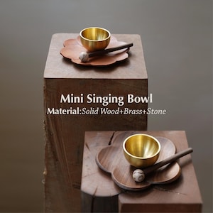 May include: A mini singing bowl set made of solid wood, brass, and stone. The set includes a small gold-colored bowl, a wooden mallet with a stone head, and a wooden flower-shaped base. The text "Mini Singing Bowl" and material details are visible.