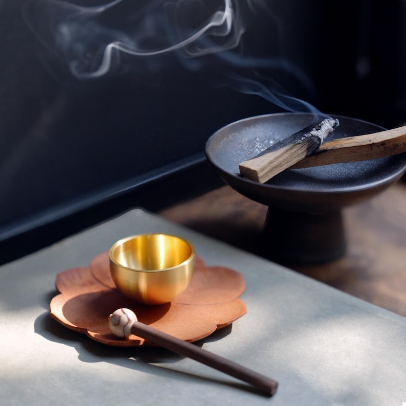 May include: A close-up of a wellness set featuring a small gold bowl, a wooden incense stick, and a dark brown incense holder with a burning incense stick. The set is arranged on a brown, flower-shaped coaster, with smoke rising in the background.