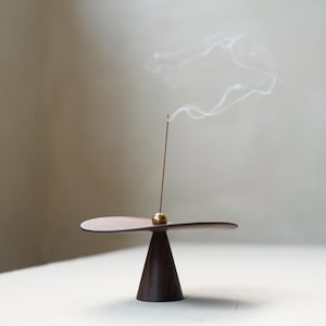 Può includere: Un porta incenso in legno marrone scuro con un accento in ottone e un bastoncino di incenso acceso che emette fumo bianco. Il supporto ha una base conica e una parte superiore piatta e ovale. Il design è minimalista ed elegante.