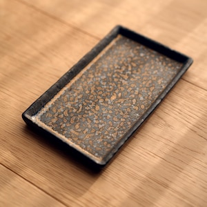 May include: Rectangular ceramic tray with a speckled, textured surface in shades of grey and gold. The tray has a dark border and is set on a wooden surface. A decorative piece for serving or display.