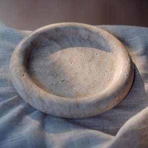 May include: A beige, circular stone tray with a shallow, concave center. The tray has a smooth, rounded rim and a textured surface. The neutral color and simple design make it suitable for various uses, such as a decorative accent or a catch-all for small items.