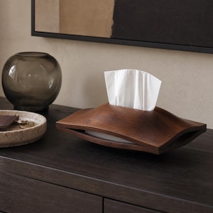 Pode incluir: Uma caixa de madeira castanho-escura para lenços com uma silhueta suave, curva e ondulada. O suporte retangular apresenta uma abertura central no topo por onde sai um lenço de papel branco, exibindo um acabamento de grão de madeira.