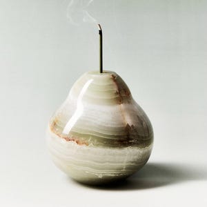 May include: A pear-shaped incense holder crafted from layered, polished stone. The holder is predominantly light green with bands of white and brown. A lit incense stick is inserted into the top, with smoke gently rising.