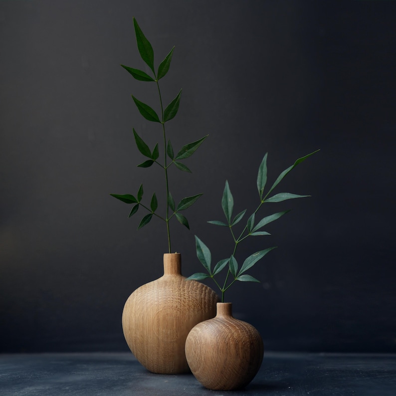 Puede incluir: Dos jarrones de madera de diferentes tama&ntilde;os, cada uno con una ramita de hojas verdes. Los jarrones tienen un acabado de vetas de madera natural y una forma redondeada. El fondo es gris oscuro.