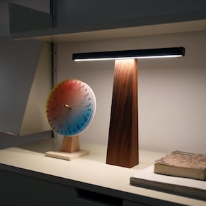 Op de afbeelding: Een moderne bureaulamp met een rechthoekige zwarte lamp en een donkere houten voet. Links van de lamp staat een kleurrijke ronde kalender met een houten standaard. Rechts ligt een boek.