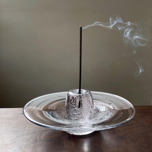 Peut inclure: Un porte-encens en verre transparent avec une coupelle centrale remplie de bulles et un plat large et peu profond. Un bâton d'encens allumé dégage une volute de fumée. Le support est posé sur une surface en bois sombre, avec un fond neutre.