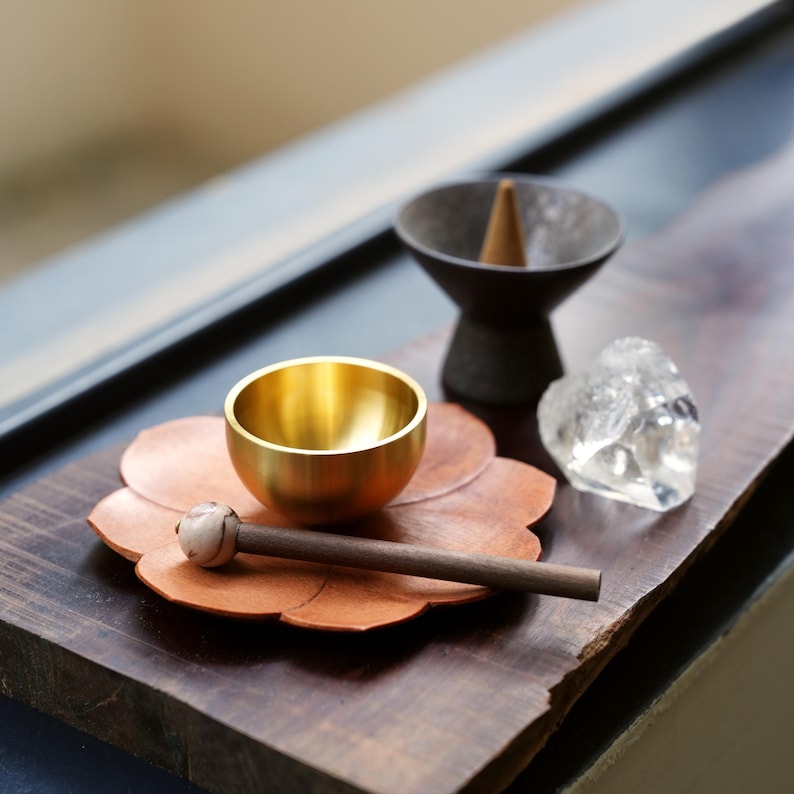 May include: A collection of items for meditation or relaxation. Includes a small, gold-colored bowl, a copper-colored flower-shaped plate, a wooden mallet, a black incense holder with a cone, and a clear crystal. All items are arranged on a dark wooden surface.