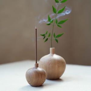 Puede incluir: Un porta incienso de madera con una varilla de incienso encendida, emitiendo humo. Junto a &eacute;l, un peque&ntilde;o jarr&oacute;n de madera contiene un tallo verde y frondoso. Los objetos est&aacute;n sobre una superficie blanca, con un fondo neutro.