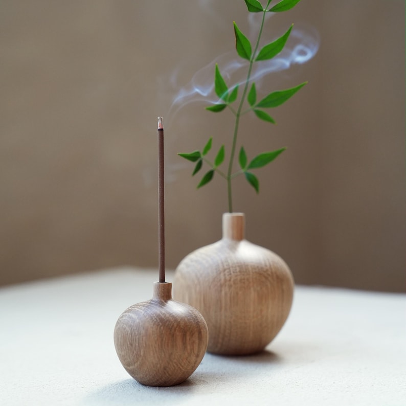Puede incluir: Un porta incienso de madera con una varilla de incienso encendida, emitiendo humo. Junto a &eacute;l, un peque&ntilde;o jarr&oacute;n de madera contiene un tallo verde y frondoso. Los objetos est&aacute;n sobre una superficie blanca, con un fondo neutro.