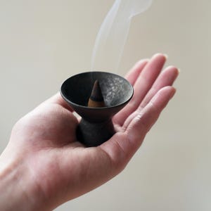 May include: A small, dark gray incense holder with a cone-shaped incense stick emitting white smoke. The holder has a flared bowl shape and a pedestal base. The incense is lit and held in an open palm against a neutral background.
