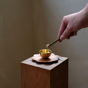 May include: A small, gold-colored bowl sits on a copper-colored flower-shaped base. A wooden mallet with a rounded head is held above the bowl. The bowl and base are on a square wooden block.