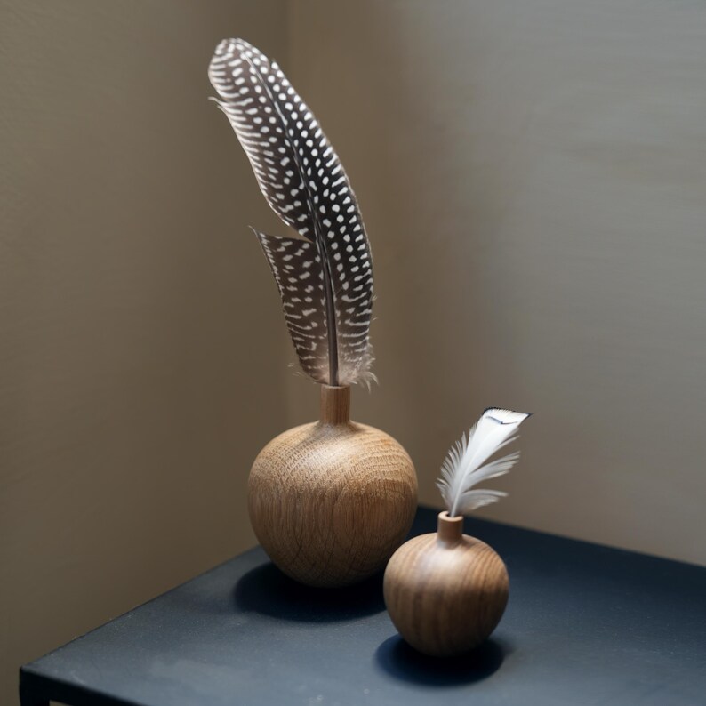 Puede incluir: Dos jarrones decorativos de madera con plumas. Uno contiene una pluma grande y moteada, y el otro una pluma blanca peque&ntilde;a. Los jarrones son esf&eacute;ricos con un cuello estrecho y est&aacute;n hechos de madera clara.