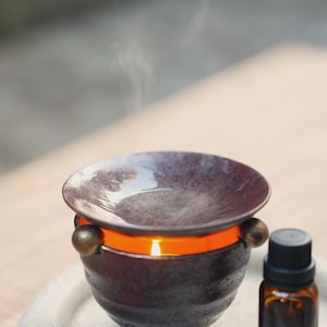 May include: A dark brown essential oil diffuser with a lit candle inside, emitting steam. The diffuser has a round, shallow dish on top and sits on a white stone tray. A small amber glass bottle with a black cap is next to it.