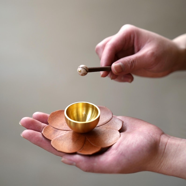 May include: A small, gold-colored singing bowl rests on a brown, flower-shaped base. A wooden mallet with a white and brown head is held above the bowl, ready to strike. The bowl and base are held in a person's hand.