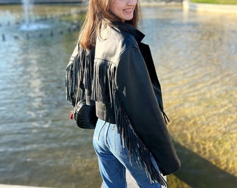 May include: A woman wearing a black leather jacket with fringe detail and blue jeans. She is standing in front of a fountain and a building with the word "fnac" on it.